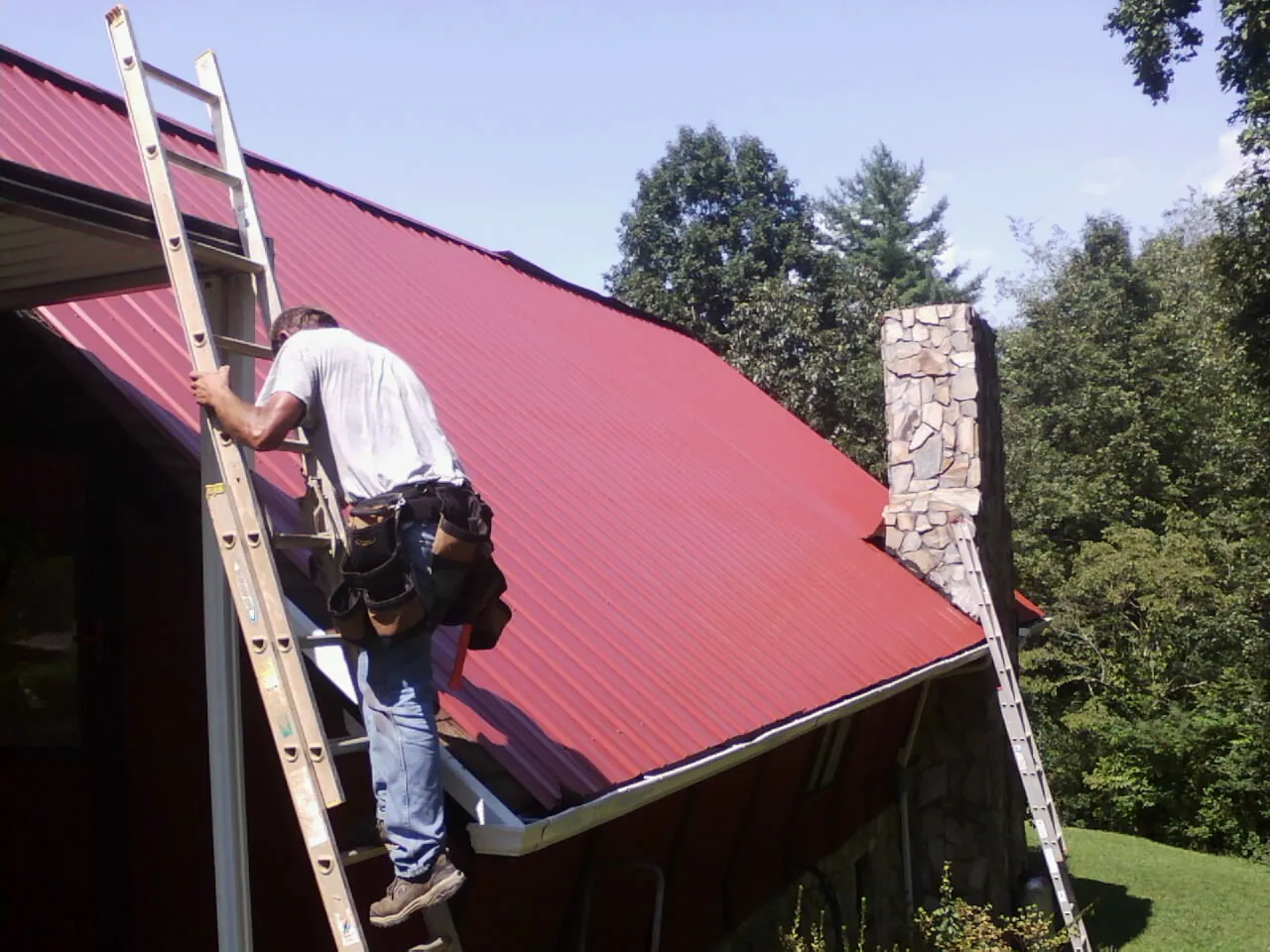 Roofer on a ladder beside a red metal roof and stone chimney — Soffit & Fascia Repair by Roofing Brothers in Ripplemead